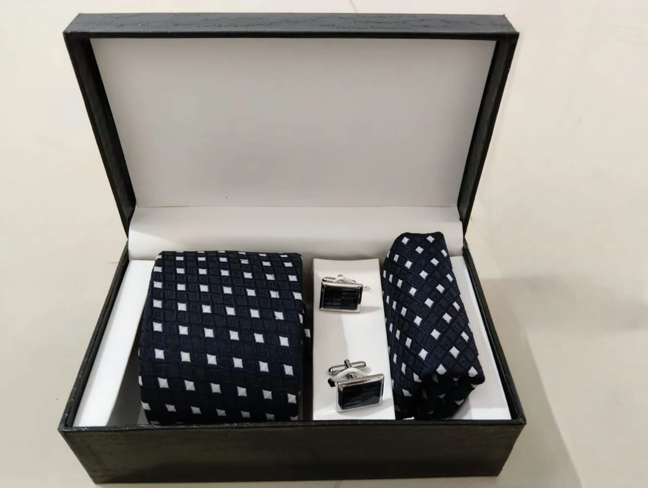 Navy blue tie with white pattern, pocket square, and cufflinks in a black box on a beige background
