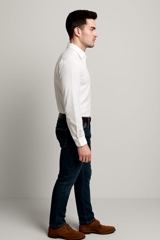 Man wearing a white dress shirt, dark jeans, and brown shoes against a plain background