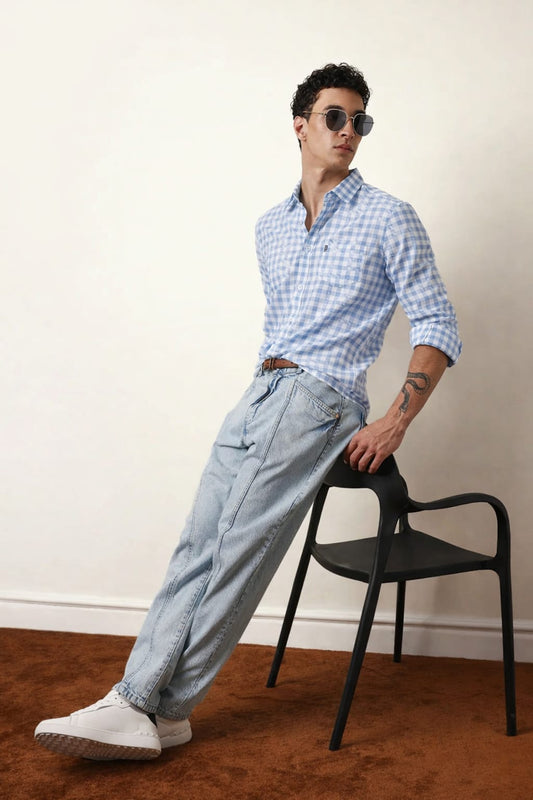 Man wearing a blue checkered shirt and light blue jeans leaning against a black chair.