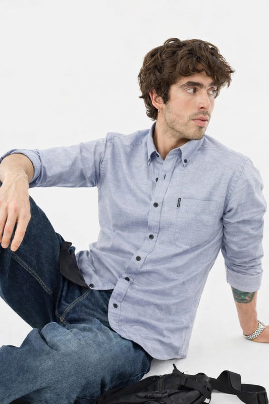 Man wearing a light blue button-up shirt and jeans sitting on a white background