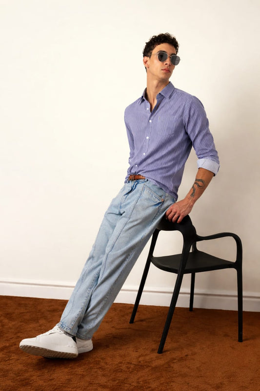 Man wearing a blue shirt and light blue jeans standing next to a black chair against a white wall.