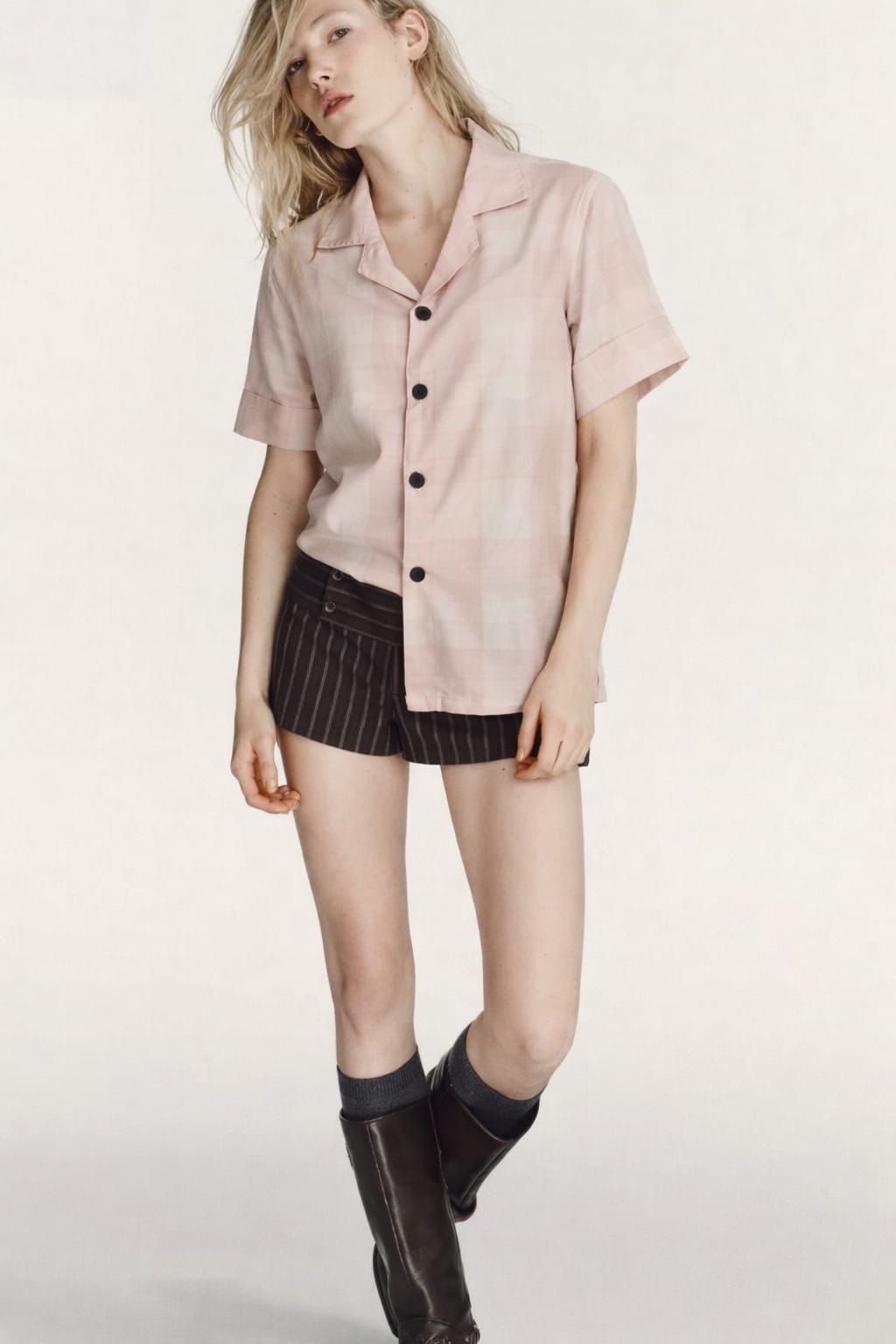 Woman wearing a light pink button-up shirt, black pinstripe skirt, and black rain boots on a white background