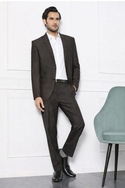 Man in a brown suit standing against a white wall with a chair in the foreground.