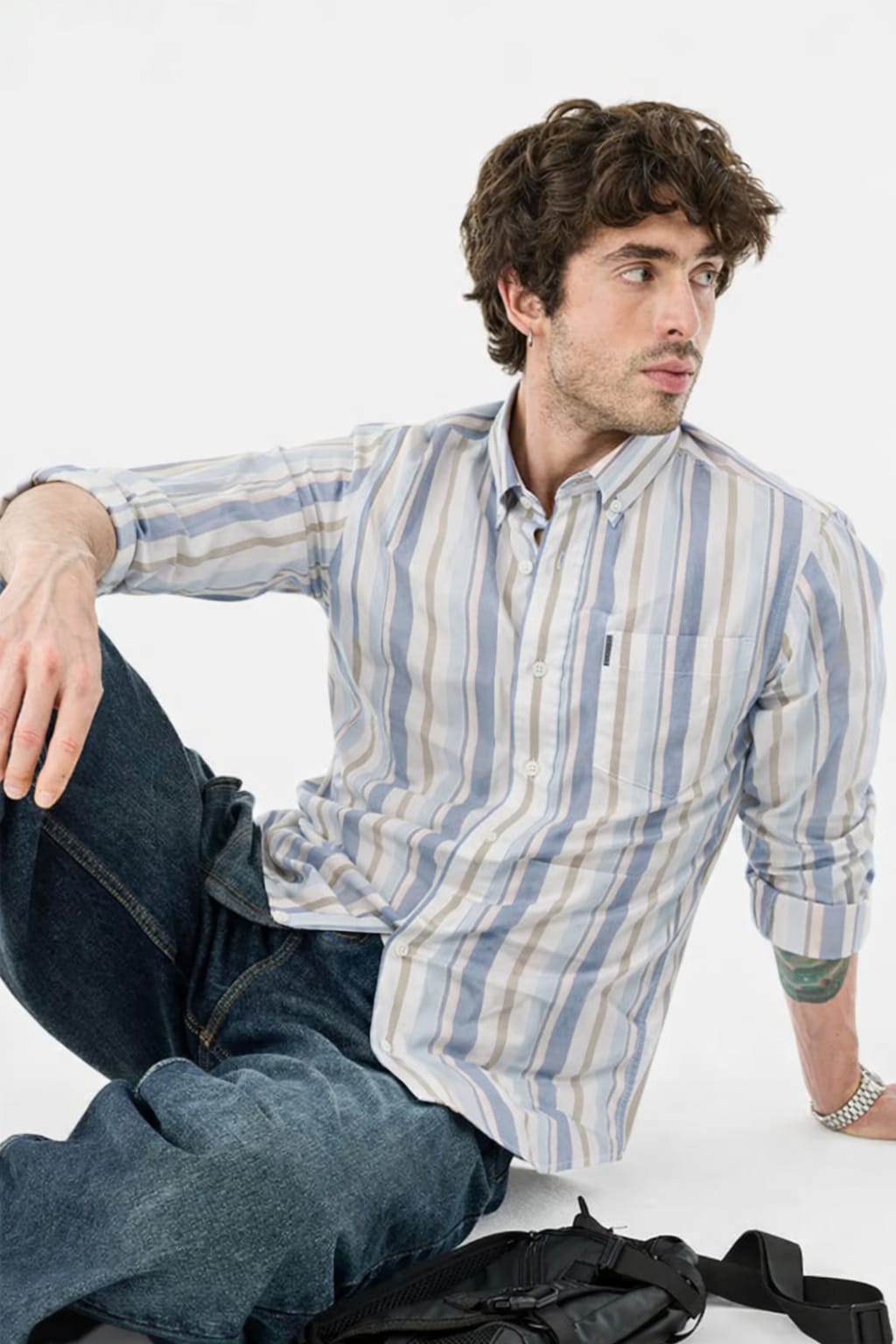 Man wearing a striped shirt and jeans sitting on the floor with a white background
