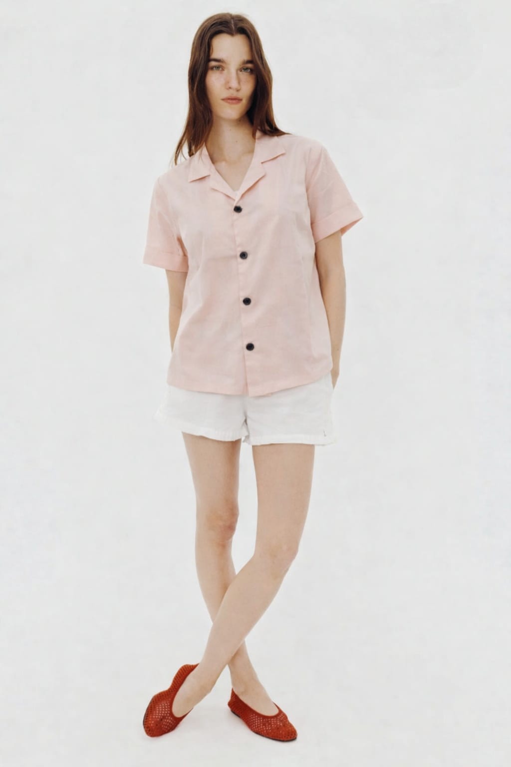 Woman wearing a light pink short-sleeve blouse with black buttons and white shorts on a white background