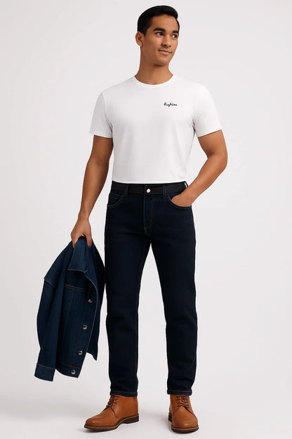 Man wearing a white t-shirt, dark jeans, and brown boots holding a navy jacket on a light gray background