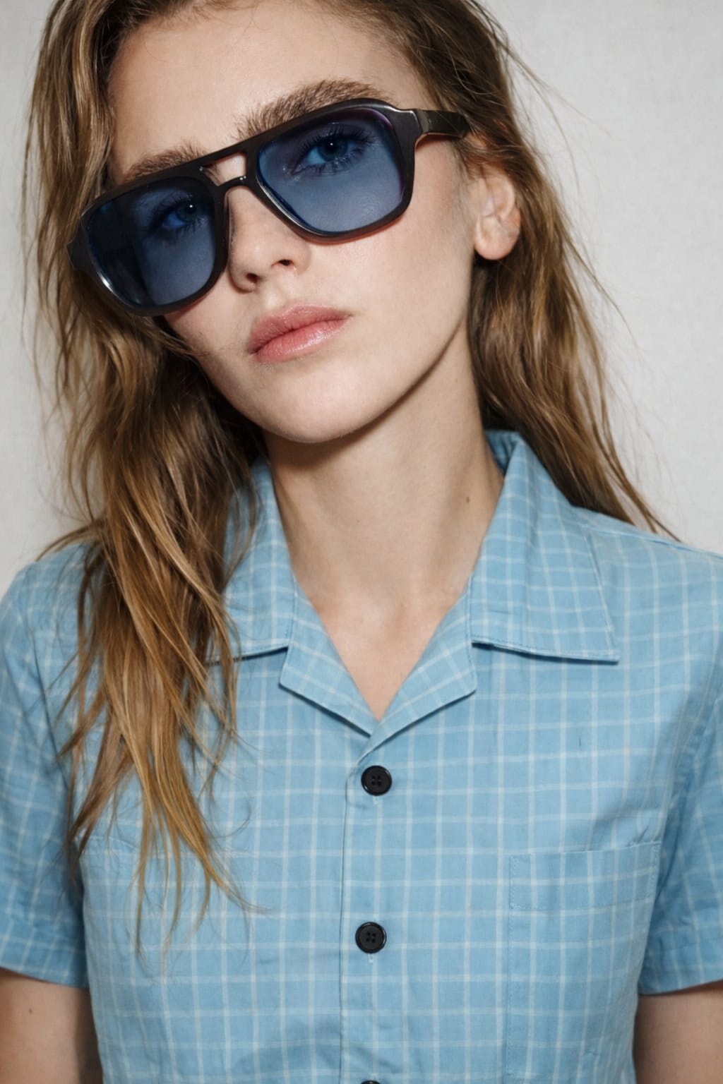 Person wearing sunglasses and a light blue checkered shirt against a neutral background