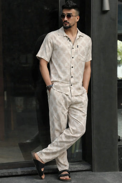 Man wearing a beige linen outfit with sunglasses, standing against a dark background.
