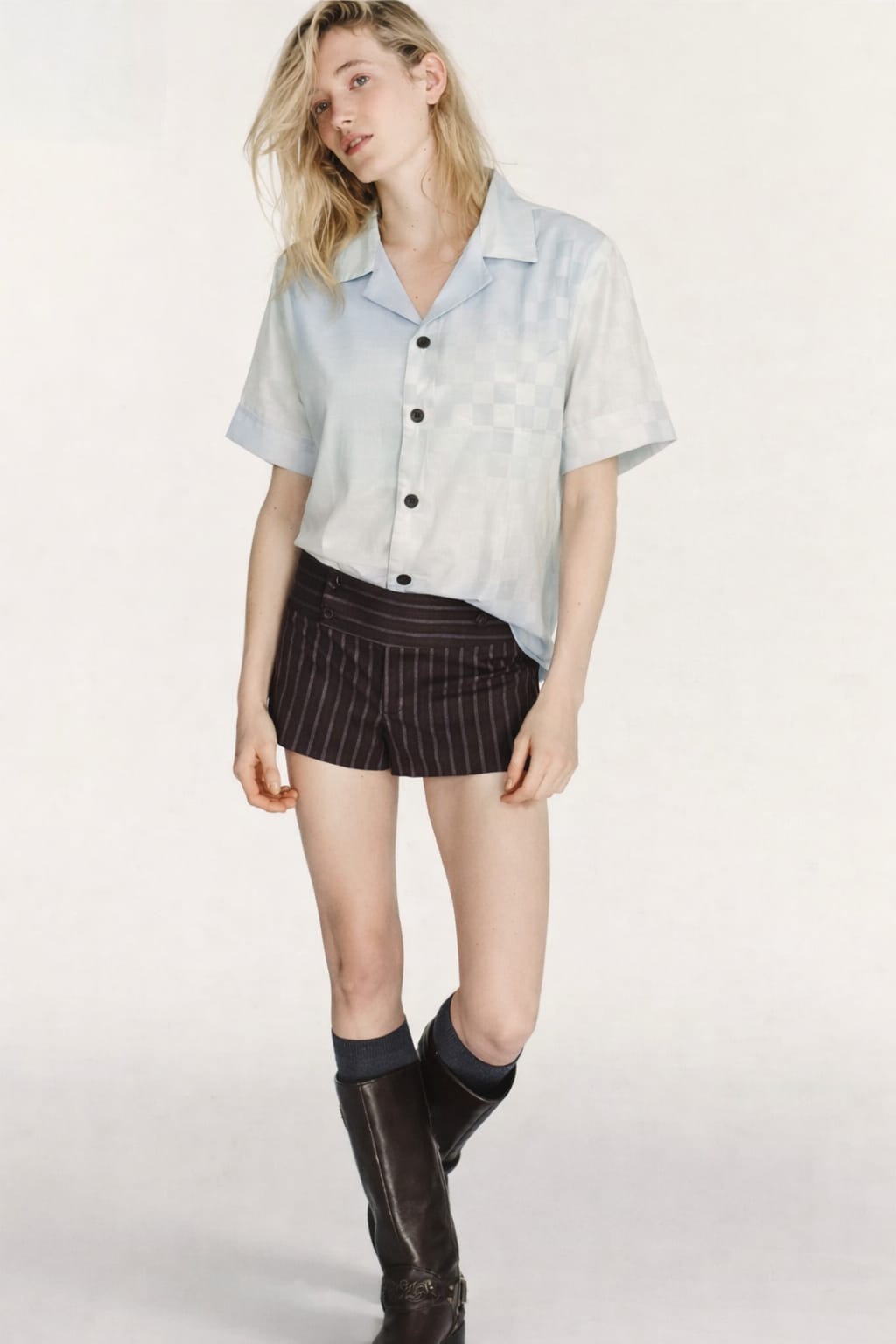 Woman wearing a light blue shirt with buttons, dark shorts, and black knee-high boots on a white background