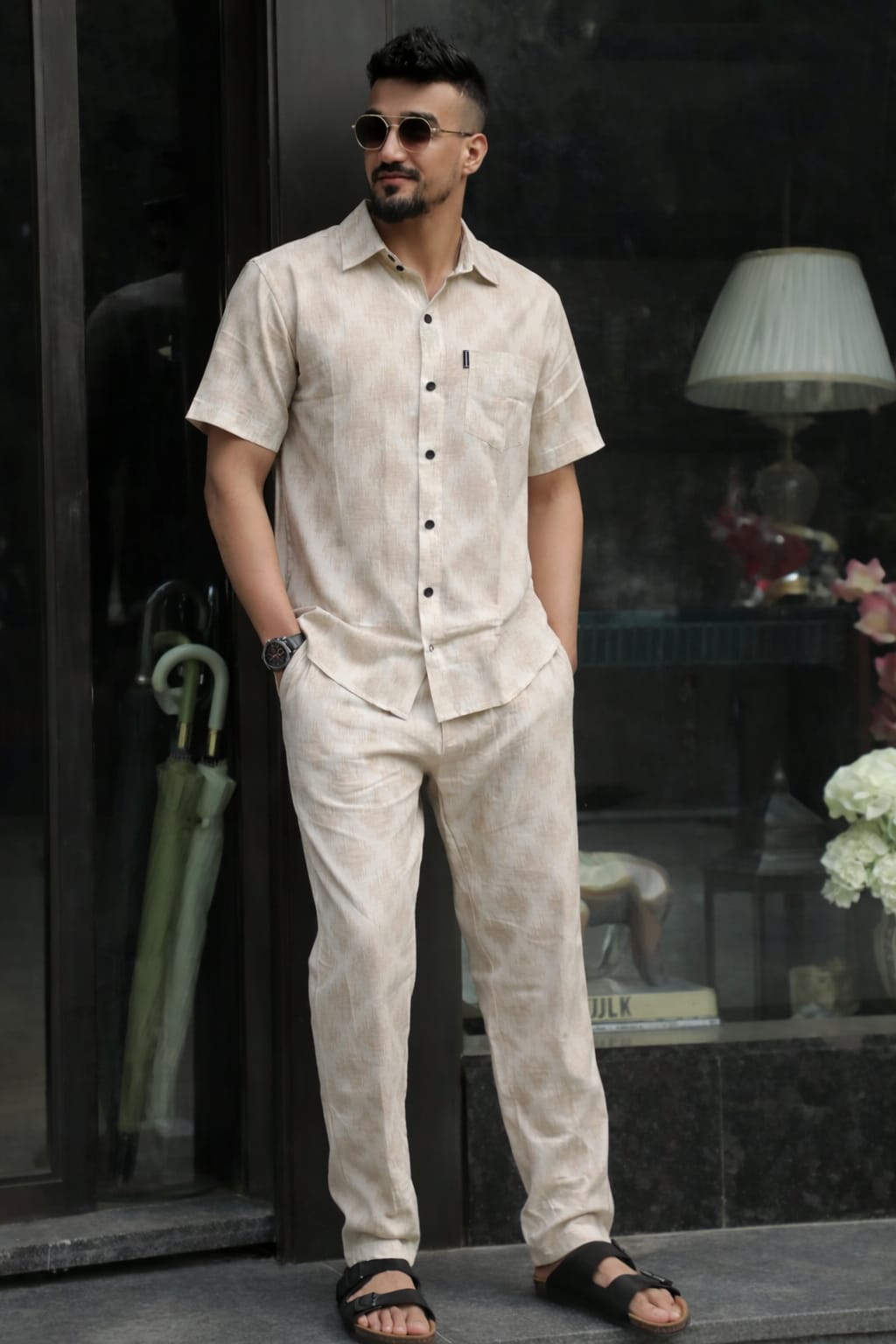 Man wearing a beige linen shirt and pants standing in front of a dark glass door.