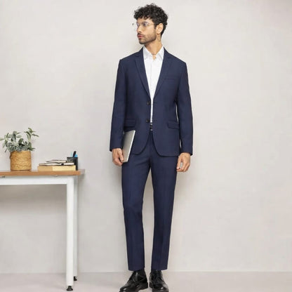 Man wearing a navy blue suit standing in a minimalistic room with a desk and plant.