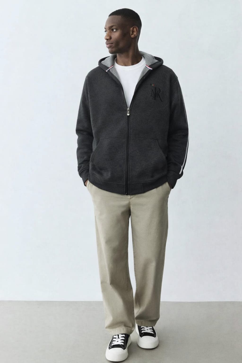 Man wearing a black jacket, white shirt, beige pants, and black sneakers against a white background
