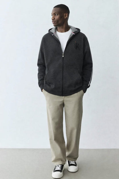 Man wearing a black jacket, white shirt, beige pants, and black sneakers against a white background