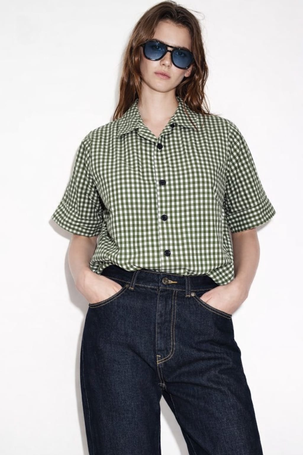 Person wearing a green checkered shirt and blue jeans with sunglasses on a white background