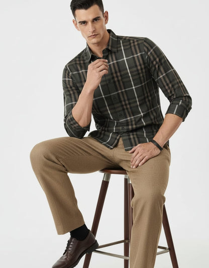 Man sitting on a stool wearing a plaid shirt and beige pants against a white background