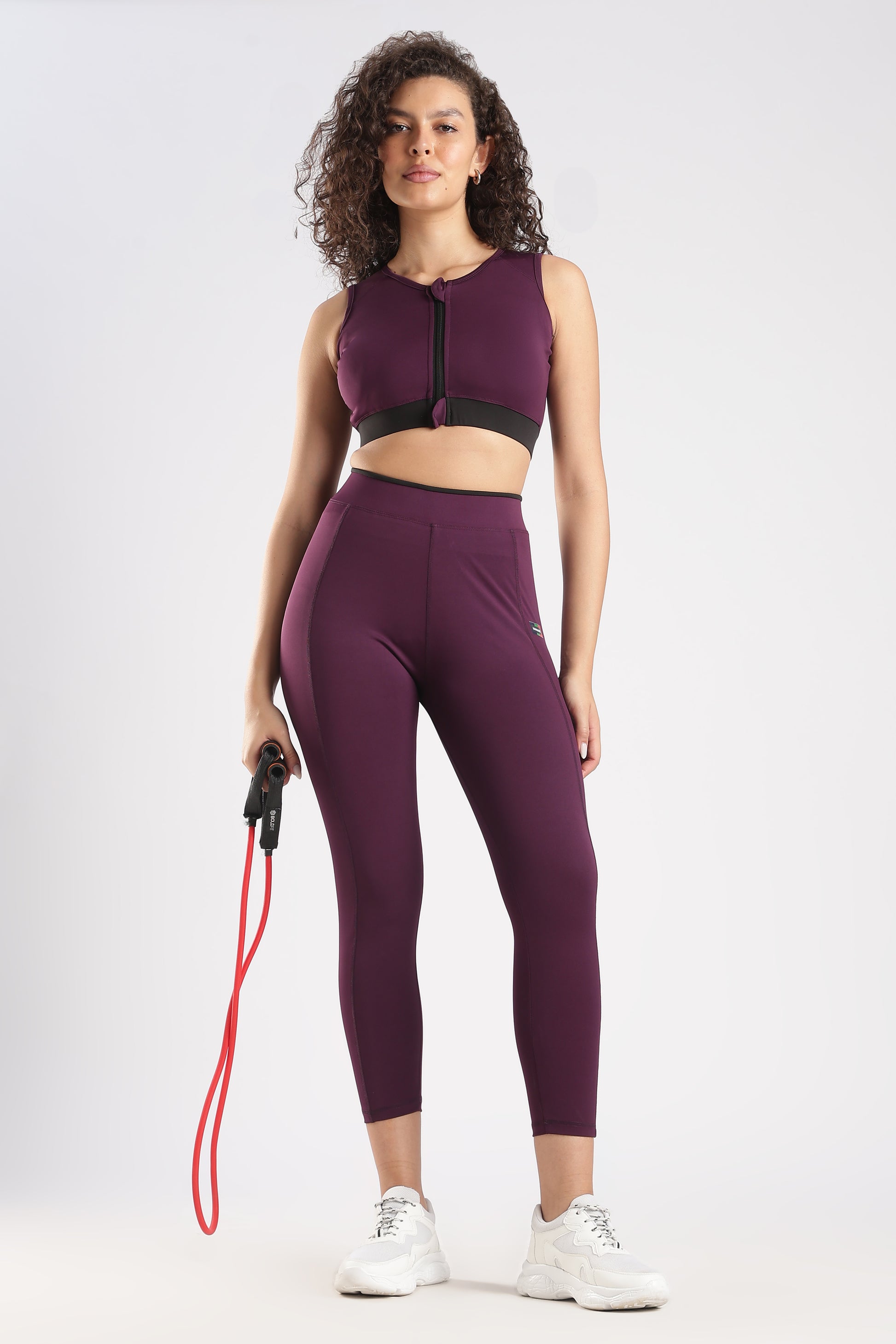 Woman wearing a purple athletic outfit with a sports bra and leggings, holding a resistance band on a white background.