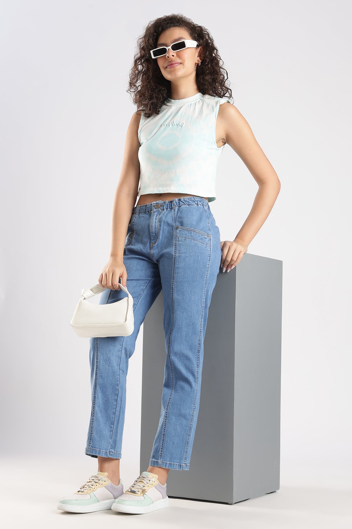Woman wearing a white sleeveless top and blue jeans, standing on a gray pedestal against a light gray background.