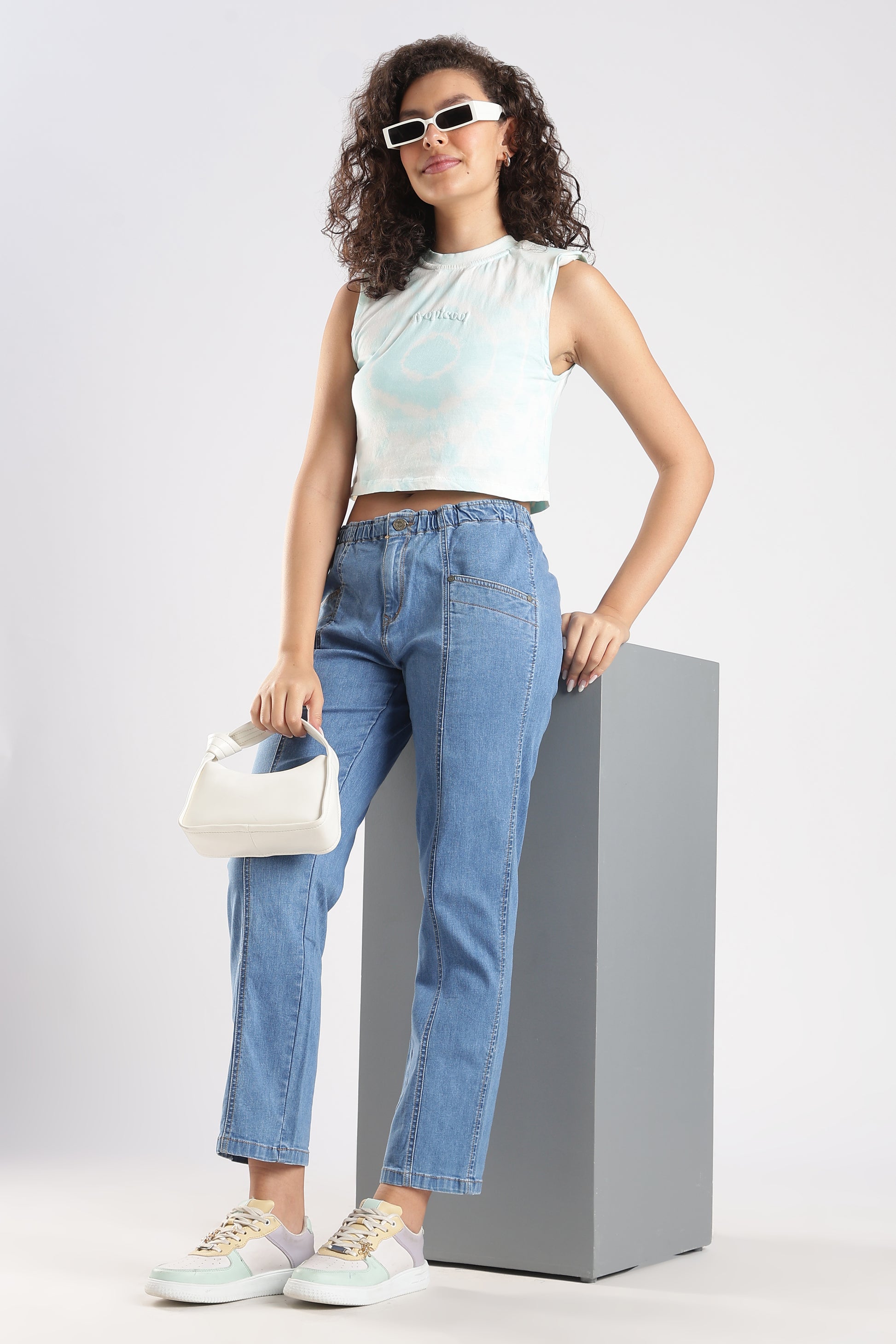 Woman wearing a white sleeveless top and blue jeans, standing on a gray pedestal against a light gray background.
