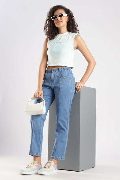 Woman wearing a white sleeveless top and blue jeans, standing on a gray pedestal against a light gray background.