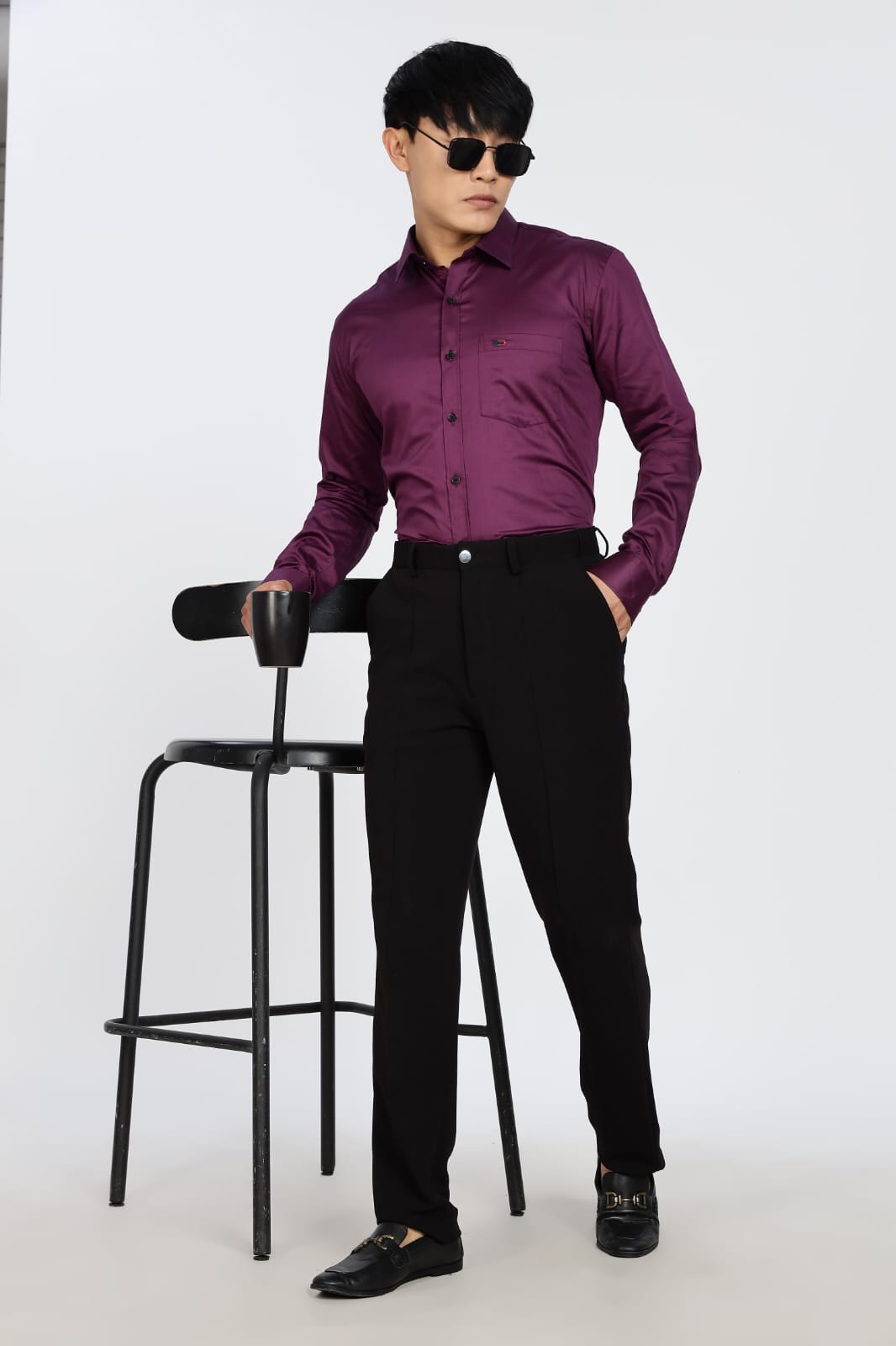 Man wearing a purple shirt and black pants standing next to a black stool on a white background
