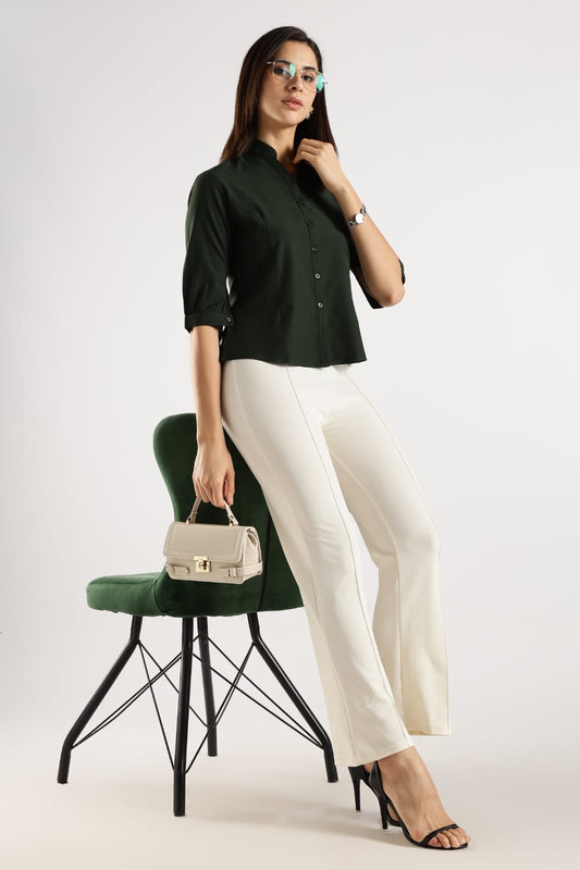 Woman in a green shirt and white pants holding a beige handbag, standing next to a green chair on a white background.