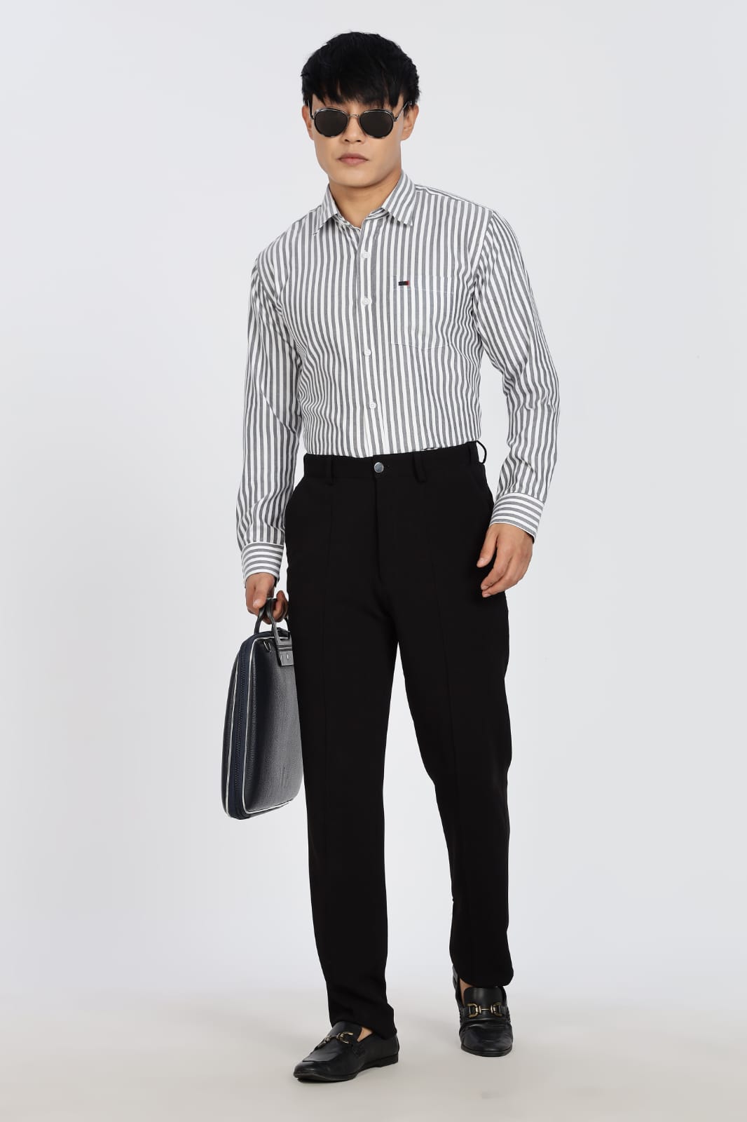 Man in a striped shirt and black pants standing next to a black stool on a white background