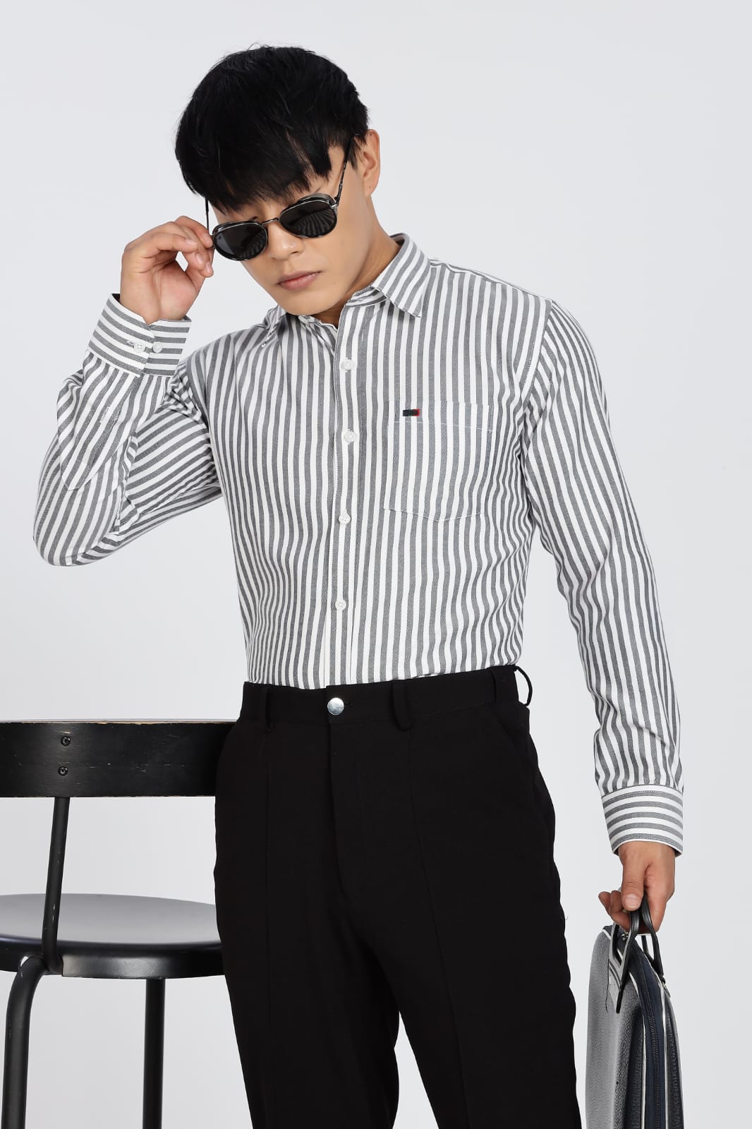 Man in a striped shirt and black pants standing next to a black stool on a white background