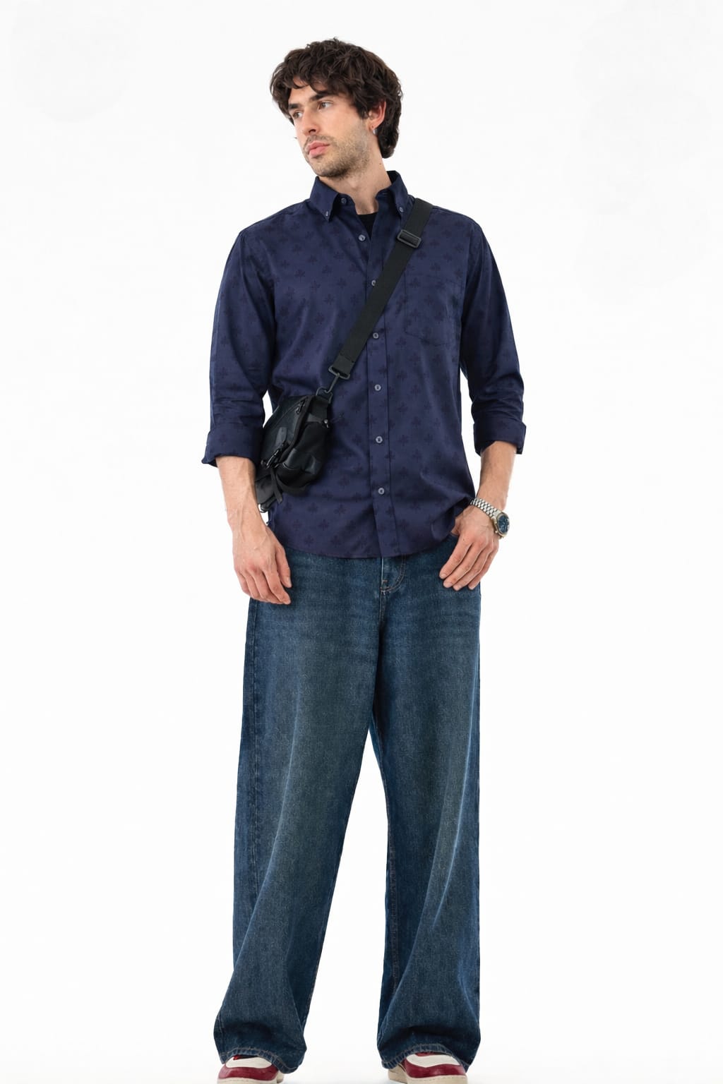 Man wearing a navy shirt and blue jeans with a black bag over one shoulder on a white background