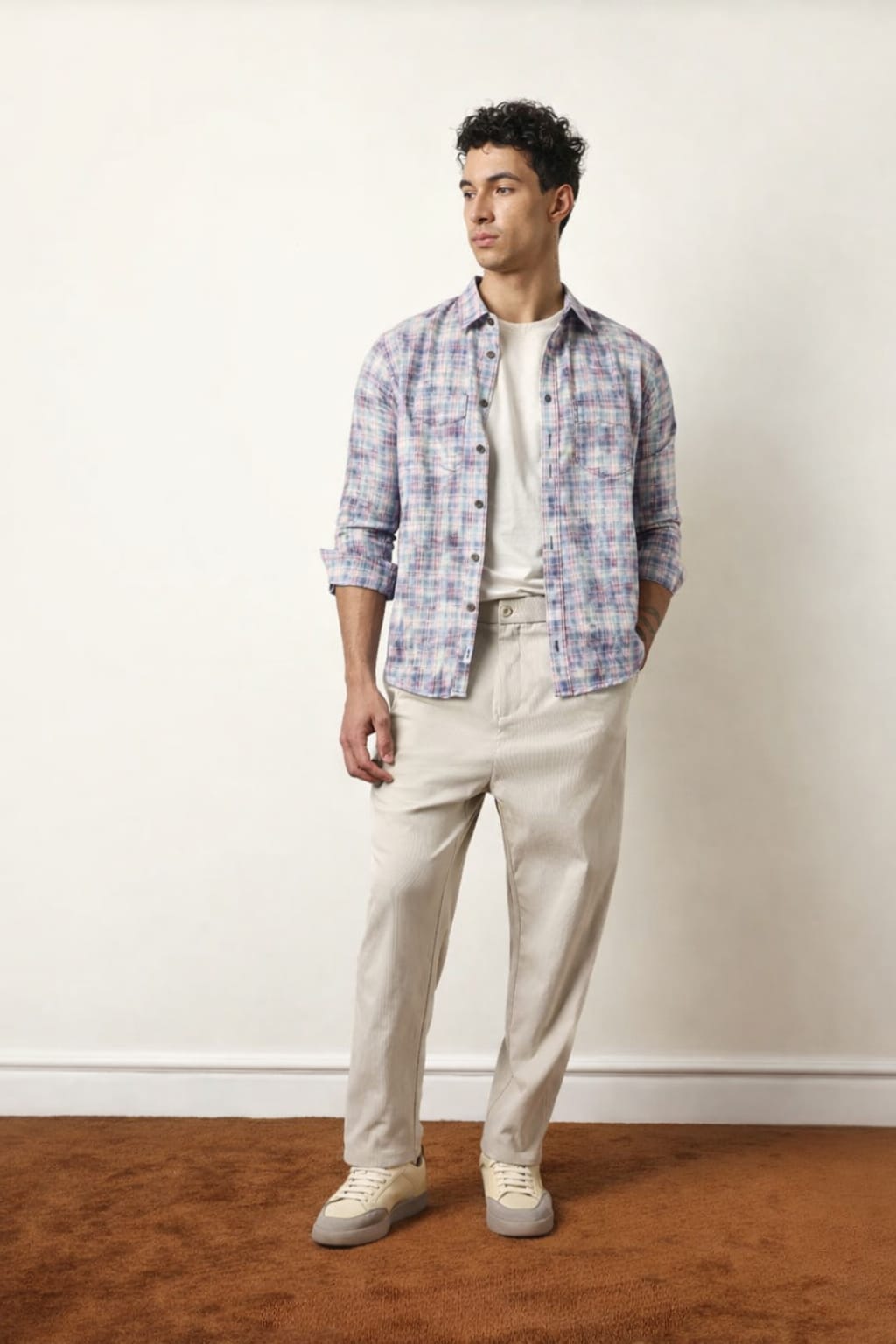Man wearing a plaid shirt, white t-shirt, beige pants, and white sneakers against a plain background.