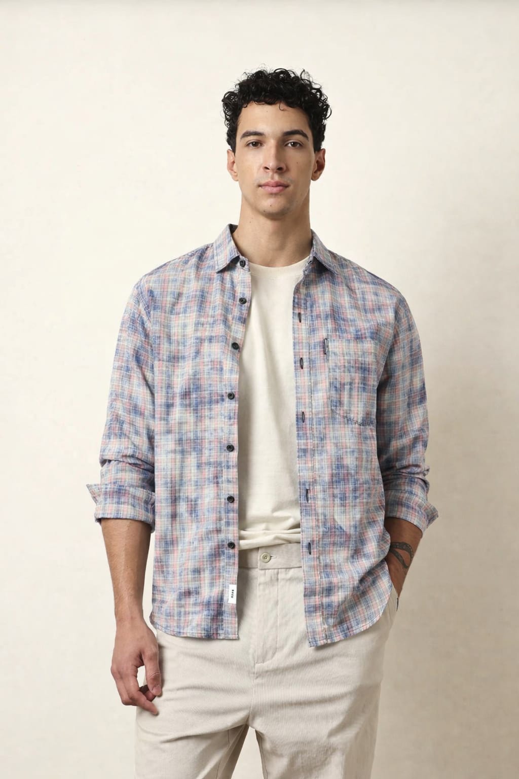 Man wearing a plaid shirt over a white t-shirt with beige pants on a plain background