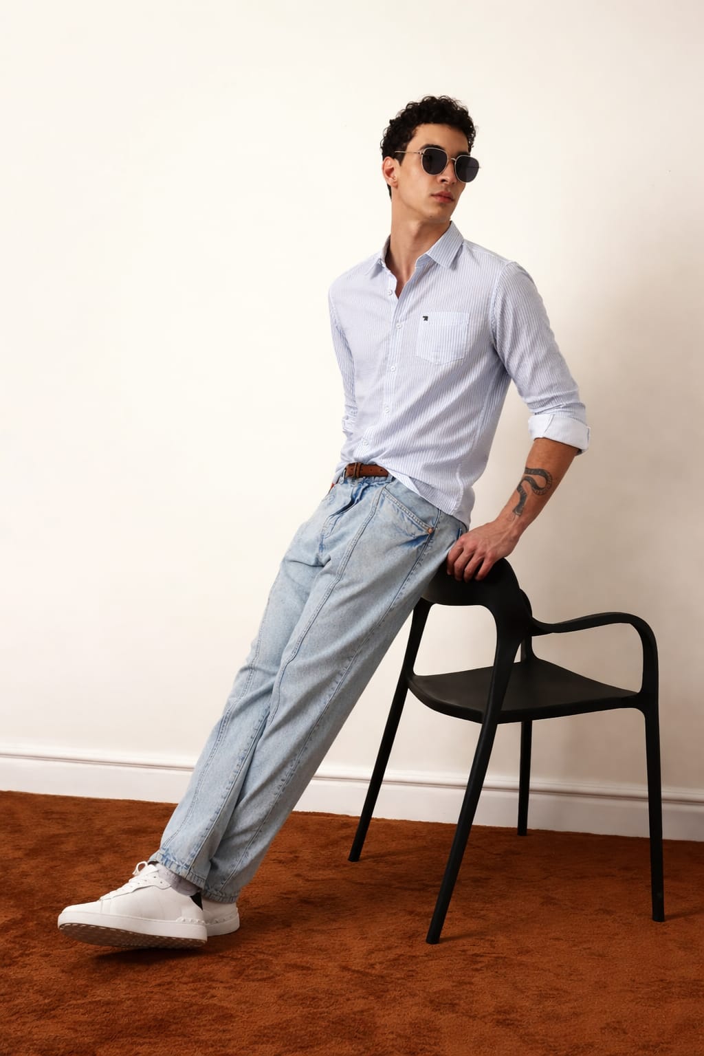 Man wearing a light blue shirt and pants sitting on a black chair against a white wall.