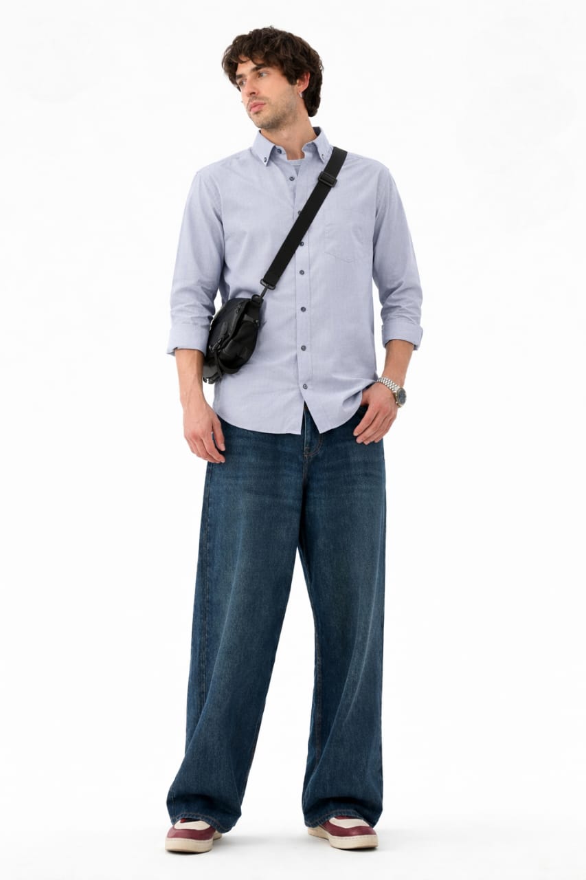 Man wearing a light blue shirt and dark jeans with a black bag over his shoulder on a white background