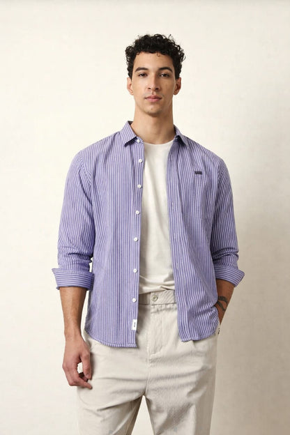 Man wearing a blue striped shirt over a white t-shirt with beige pants on a plain background