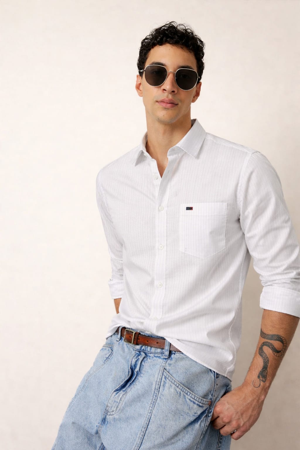 Man wearing a white shirt and light blue jeans with sunglasses on a plain background