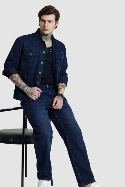 Man wearing a dark denim jacket and jeans sitting on a chair against a white background