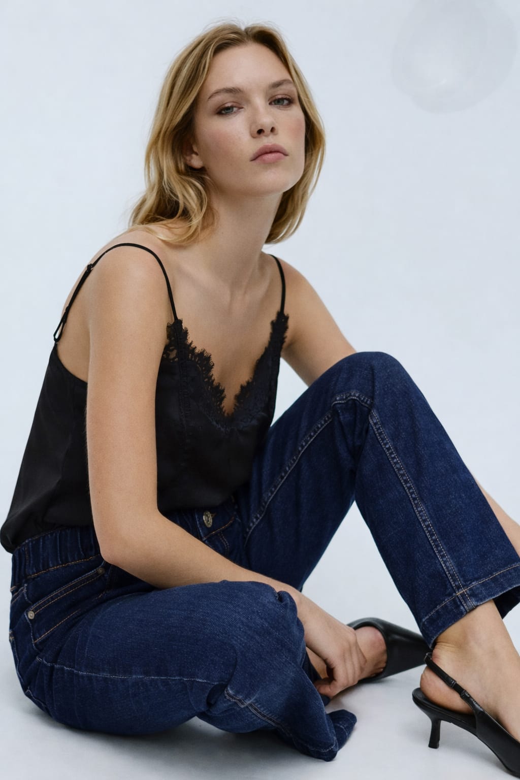 Woman wearing a black top and blue jeans sitting on a white background