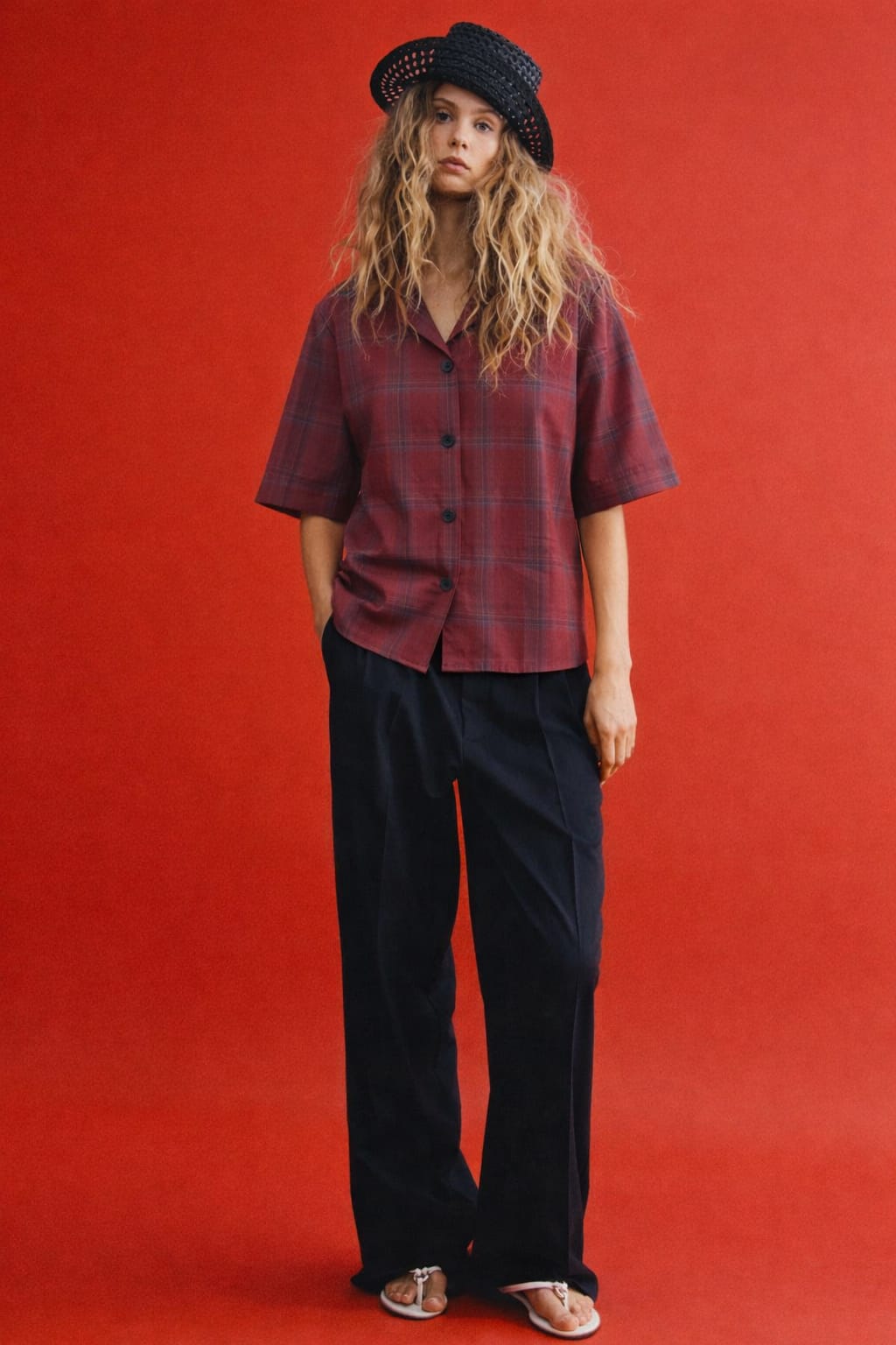 Person wearing a plaid shirt and black pants against a red background
