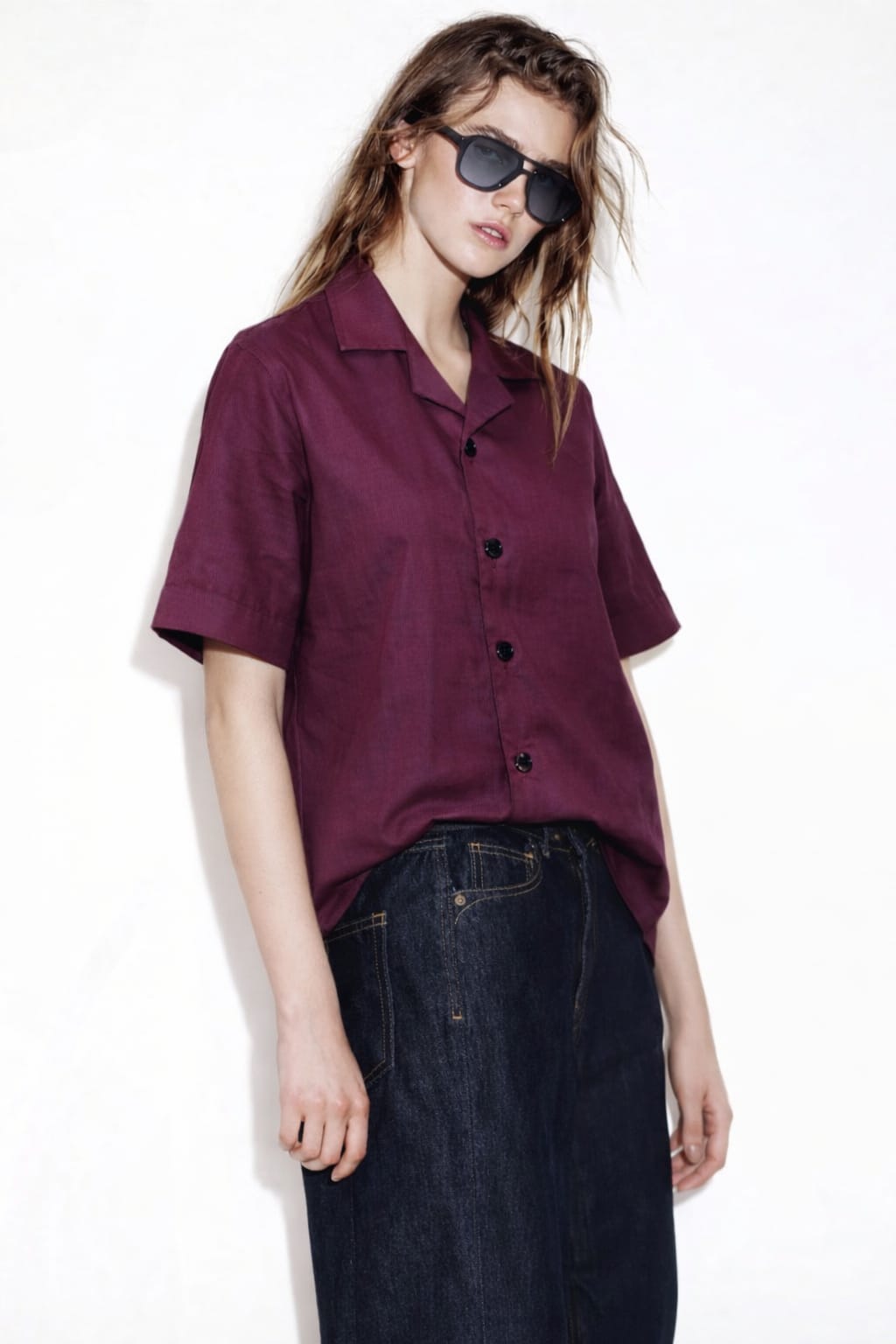 Person wearing a burgundy shirt and dark jeans with sunglasses on a white background
