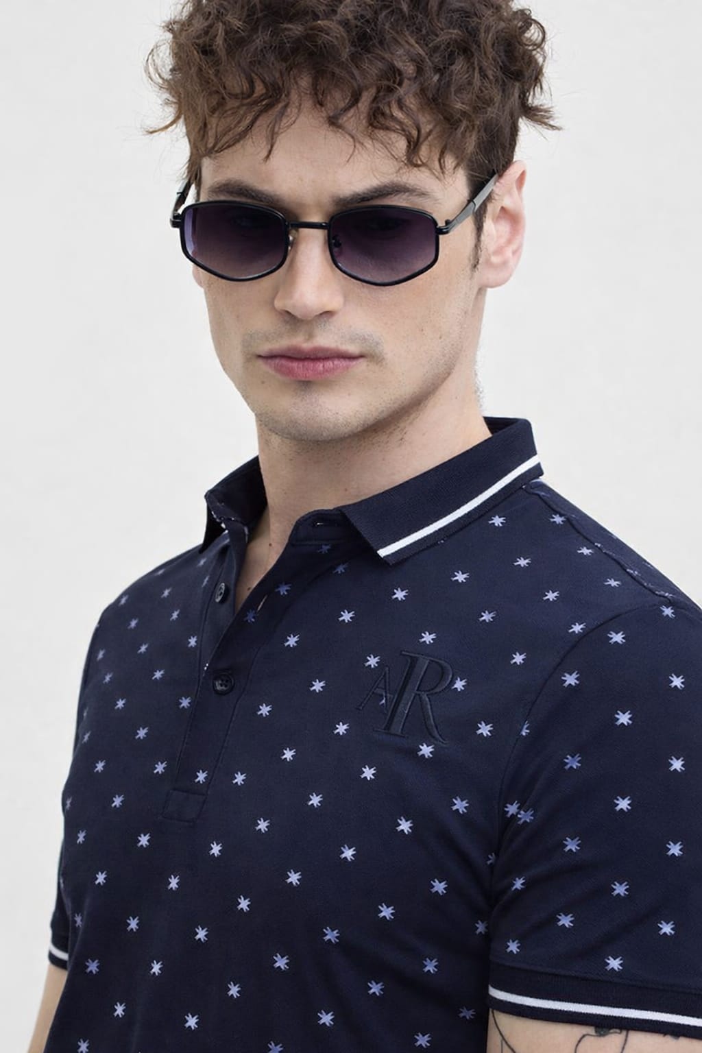 Man wearing a navy blue polo shirt with star pattern and sunglasses on a light gray background