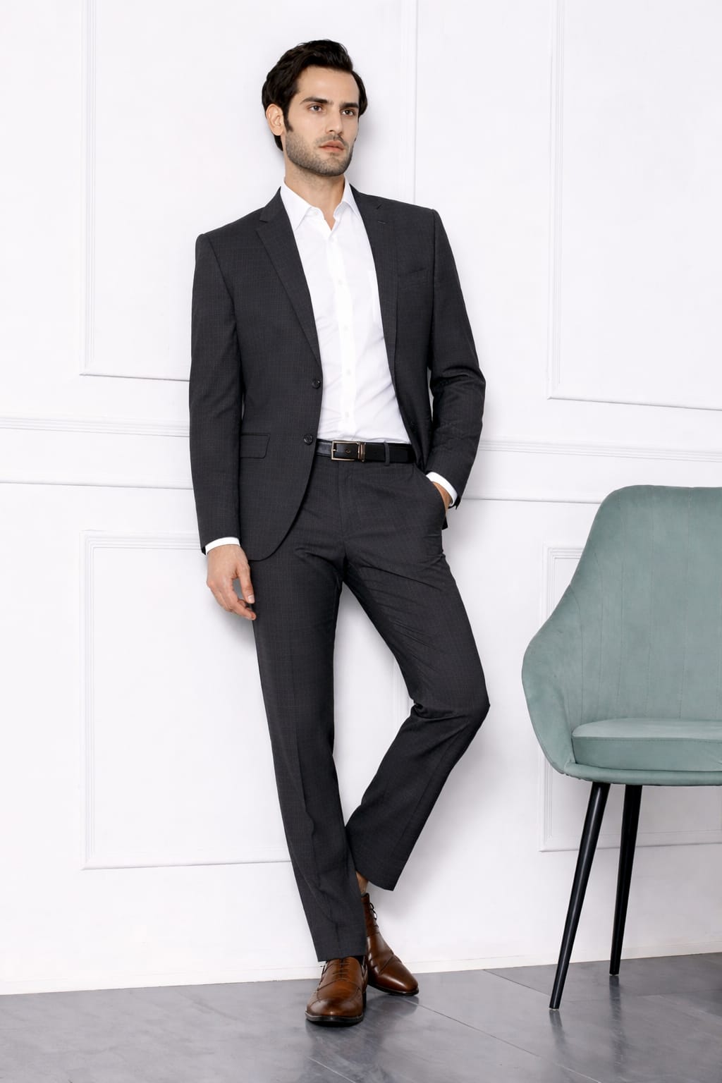 Man wearing a dark suit with a white shirt and brown shoes against a white wall.