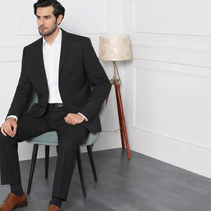 Man in a black suit sitting on a chair in a minimalistic room with a lamp in the background.
