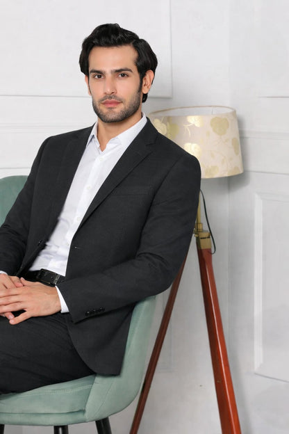 Man in a black suit sitting on a green chair with a white wall and lamp in the background