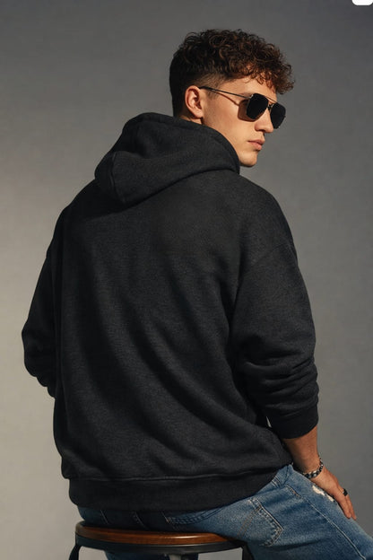 Person wearing a black hoodie and sunglasses, sitting on a stool against a gray background