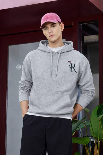 Person wearing a gray hoodie with a logo, pink cap, and black pants in front of a reflective surface.