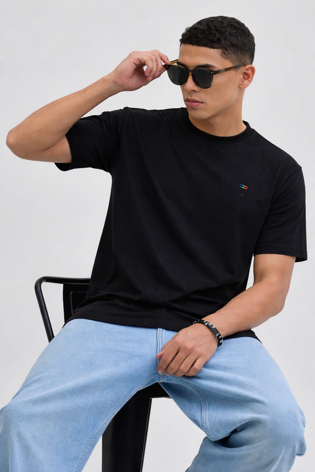 Man wearing a black t-shirt and light blue jeans sitting on a chair with sunglasses.