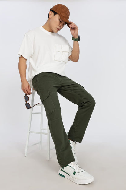 Person wearing a white shirt, green pants, and a brown cap, sitting on a stool against a white background.