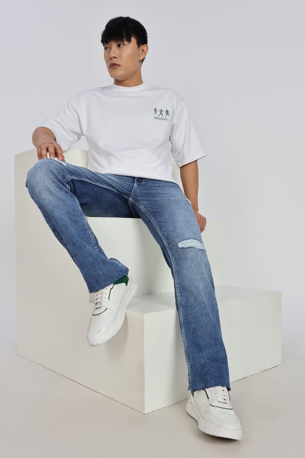 Person wearing a white t-shirt and blue jeans sitting on a white cube.