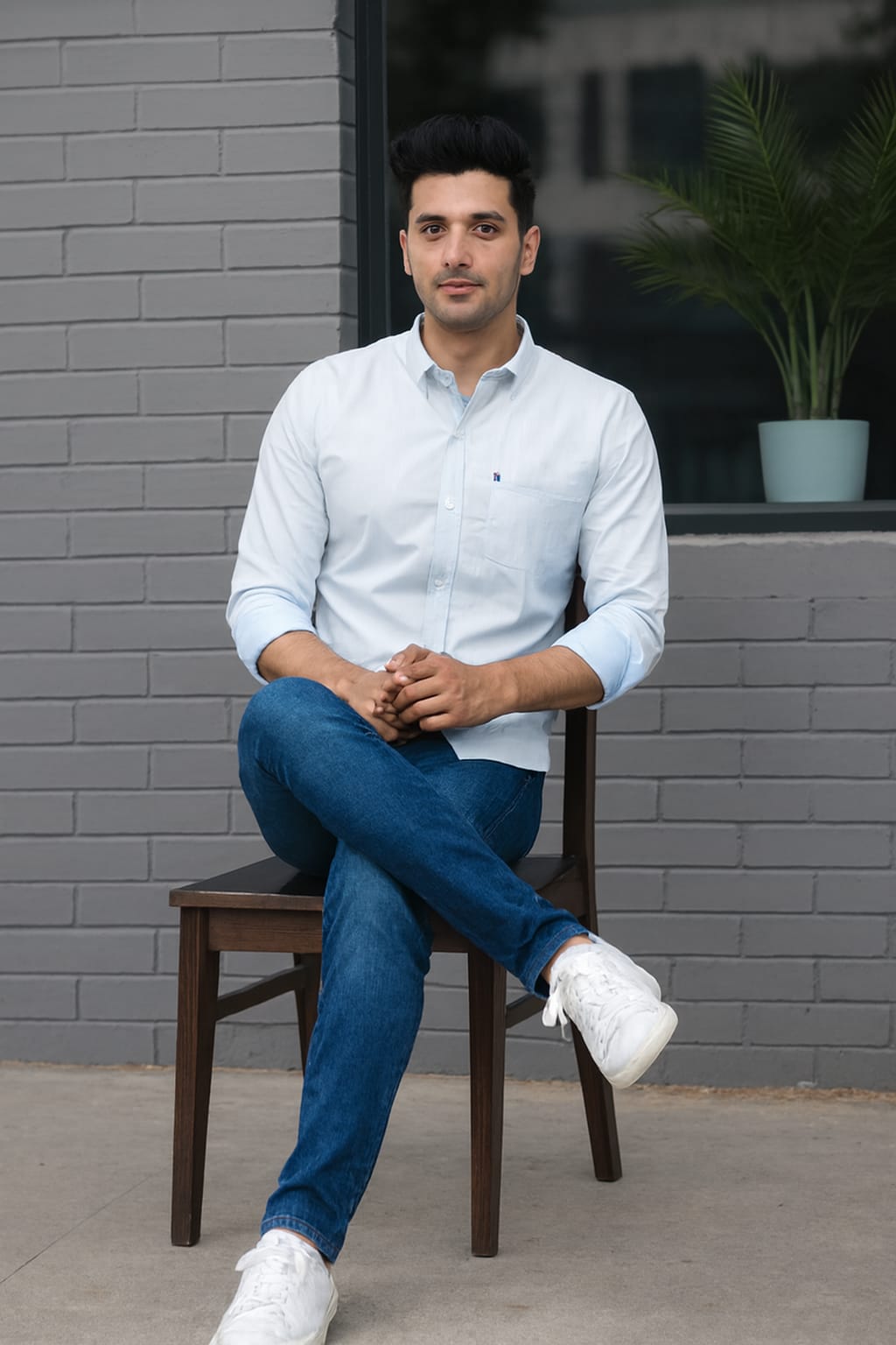 Man sitting on a chair wearing a light blue shirt and dark jeans against a gray brick wall.