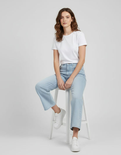 Woman wearing a white t-shirt and light blue jeans sitting on a white stool against a plain background