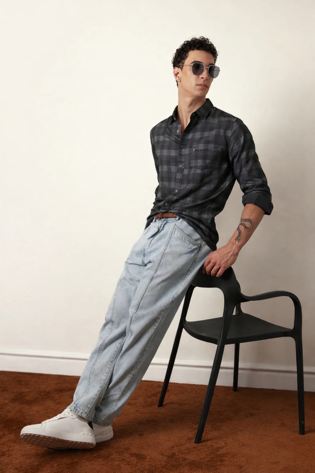 Man wearing a plaid shirt and light blue jeans leaning against a black chair.